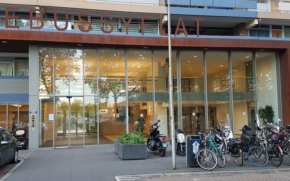 Gevel van kringloopwinkel met grote glazen puien, ingang met deuren, fietsen geparkeerd ervoor en plantenbak