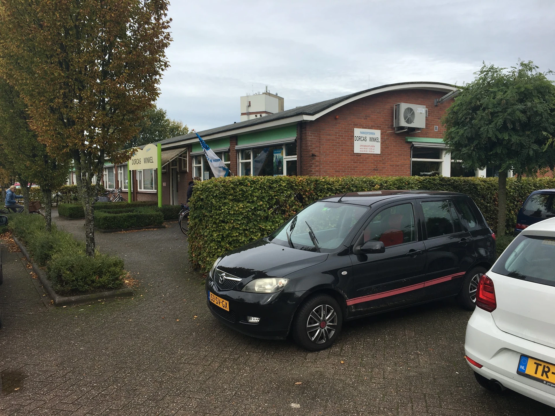 Voorgevel van een bakstenen kringloopwinkel met groene luifel, parkeerplaats met auto's ervoor