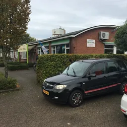 Voorgevel van een bakstenen kringloopwinkel met groene luifel, parkeerplaats met auto's ervoor