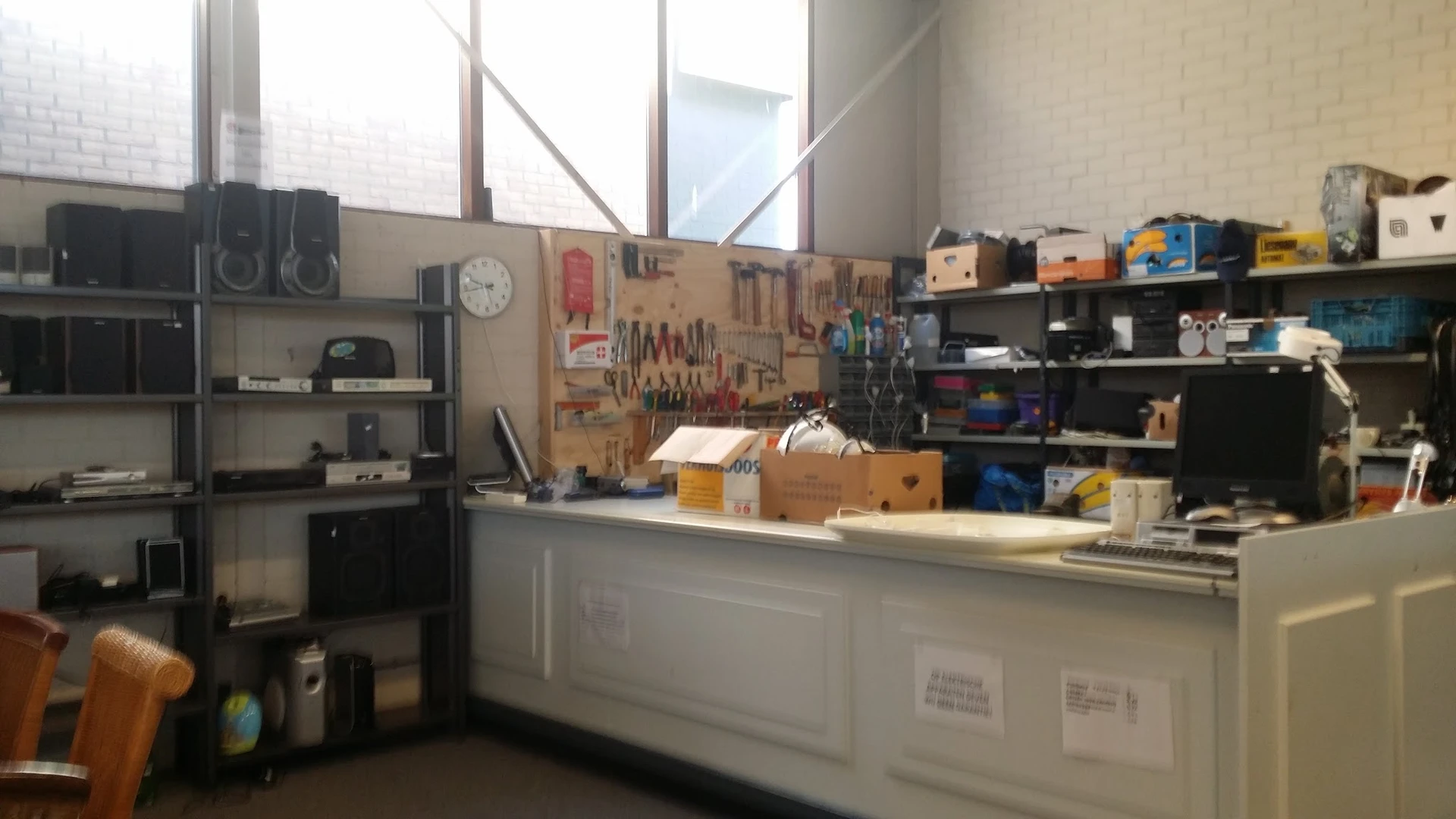 Werkruimte met gereedschapsbord, werkbank, stellingen met gereedschap en materialen, en grote ramen.