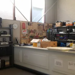 Werkruimte met gereedschapsbord, werkbank, stellingen met gereedschap en materialen, en grote ramen.