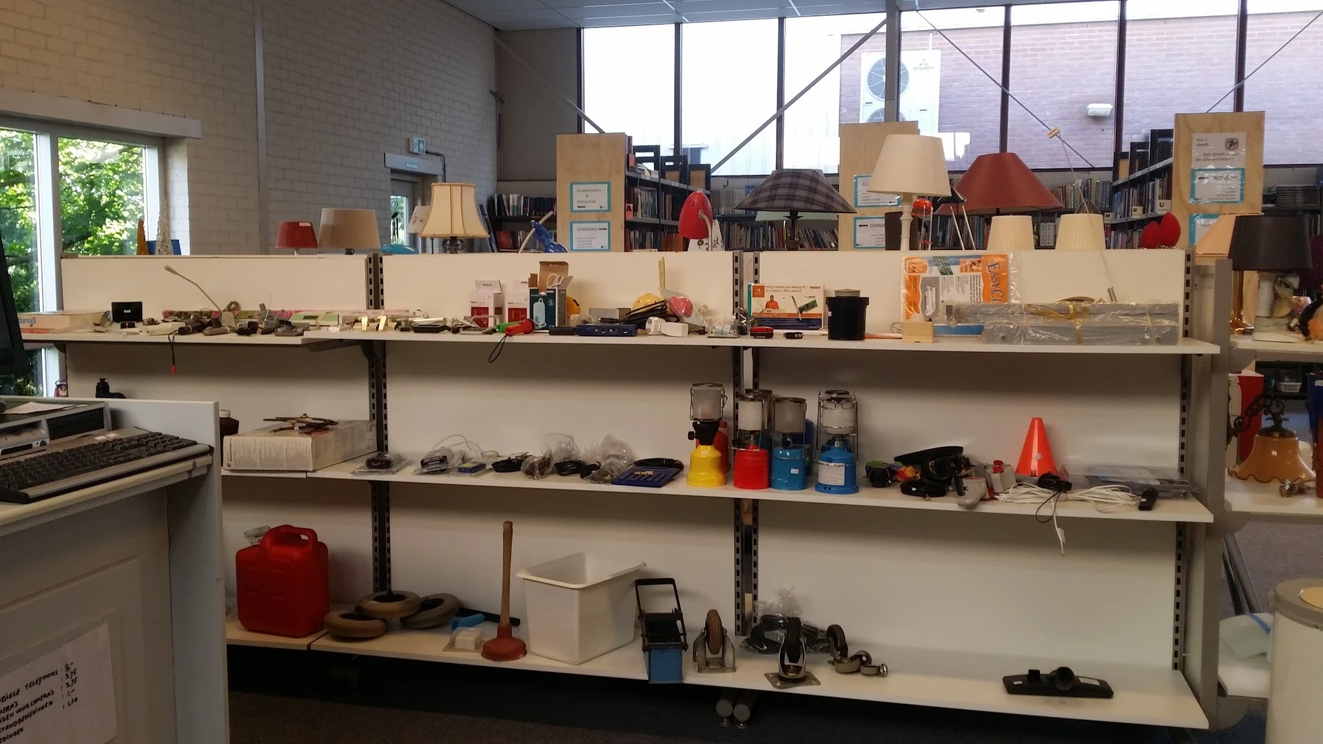 Metalen stellingen met diverse tweedehandsartikelen zoals gereedschap, huishoudspullen, lampen en dozen.