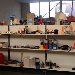 Metalen stellingen met diverse tweedehandsartikelen zoals gereedschap, huishoudspullen, lampen en dozen.