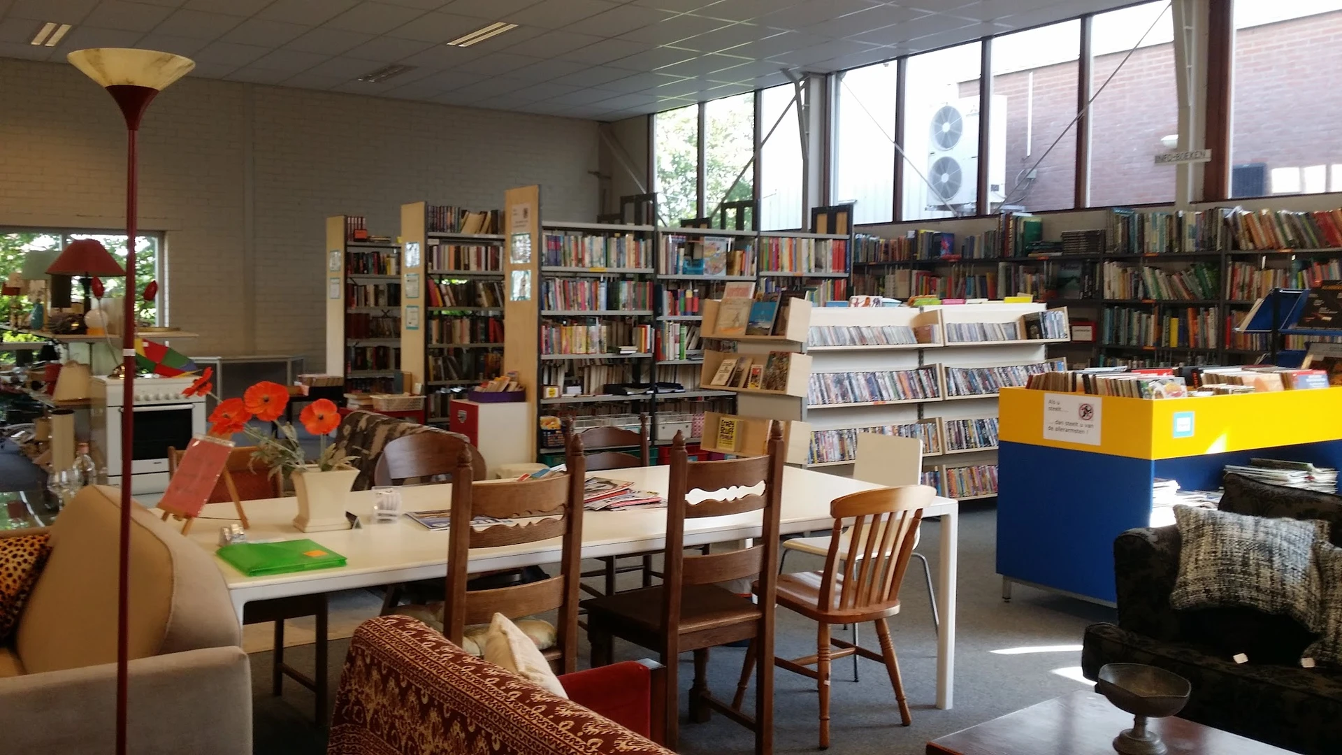 Leeshoek met houten stoelen rond een tafel, boekenkasten en media-uitstalling in kringloopwinkel