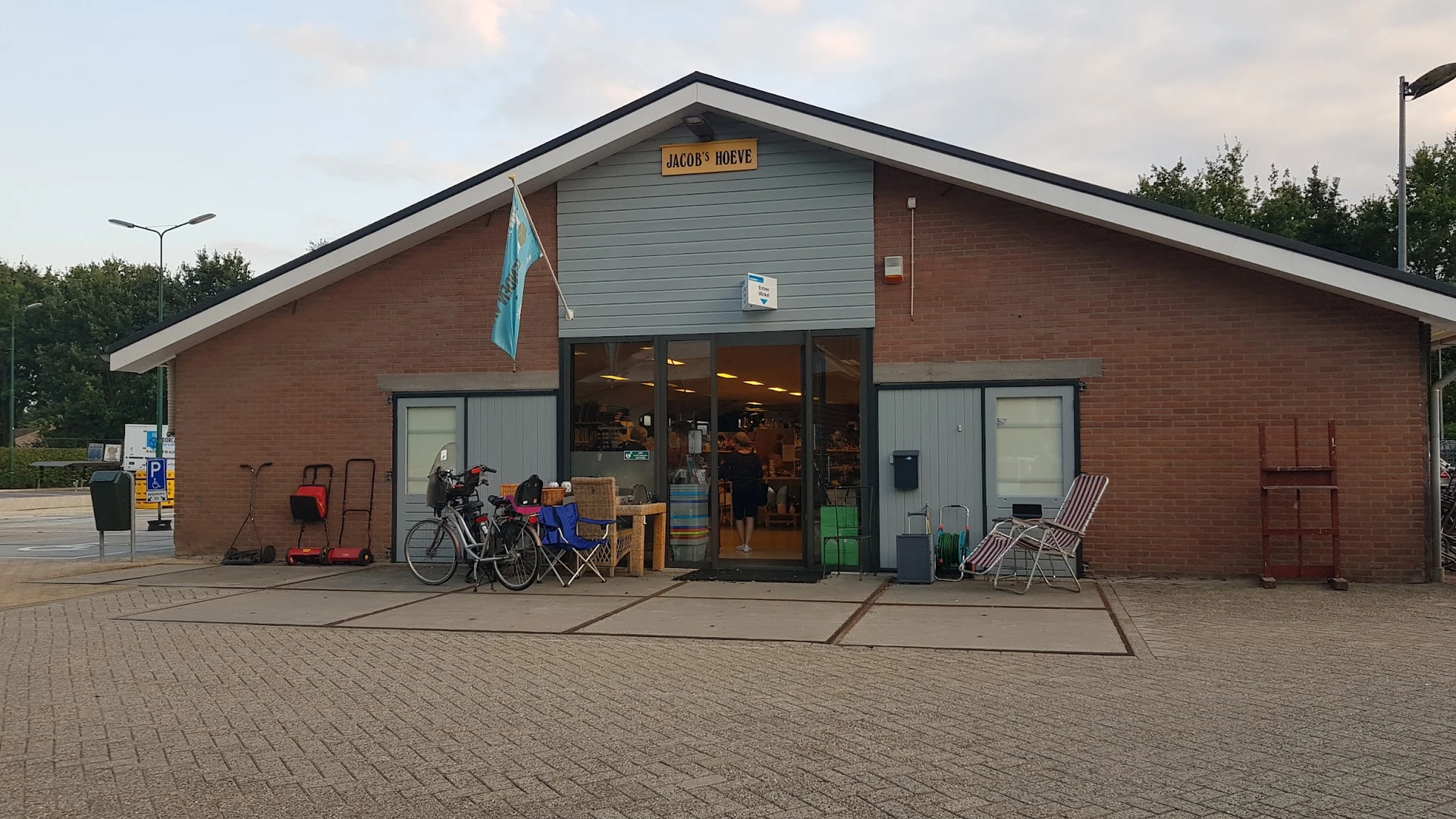 Voorkant van een bakstenen kringloopwinkel met glazen deuren, fietsenstalling en stoelen buiten