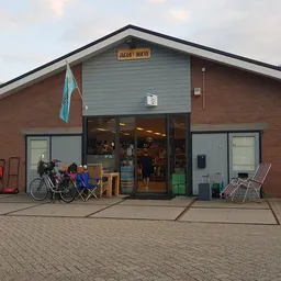 Voorkant van een bakstenen kringloopwinkel met glazen deuren, fietsenstalling en stoelen buiten