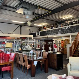 Ruime winkelvloer met donkerhouten eettafel, stoelen, rode accessoires, glazen vitrine, stellingen met spullen en houten trap
