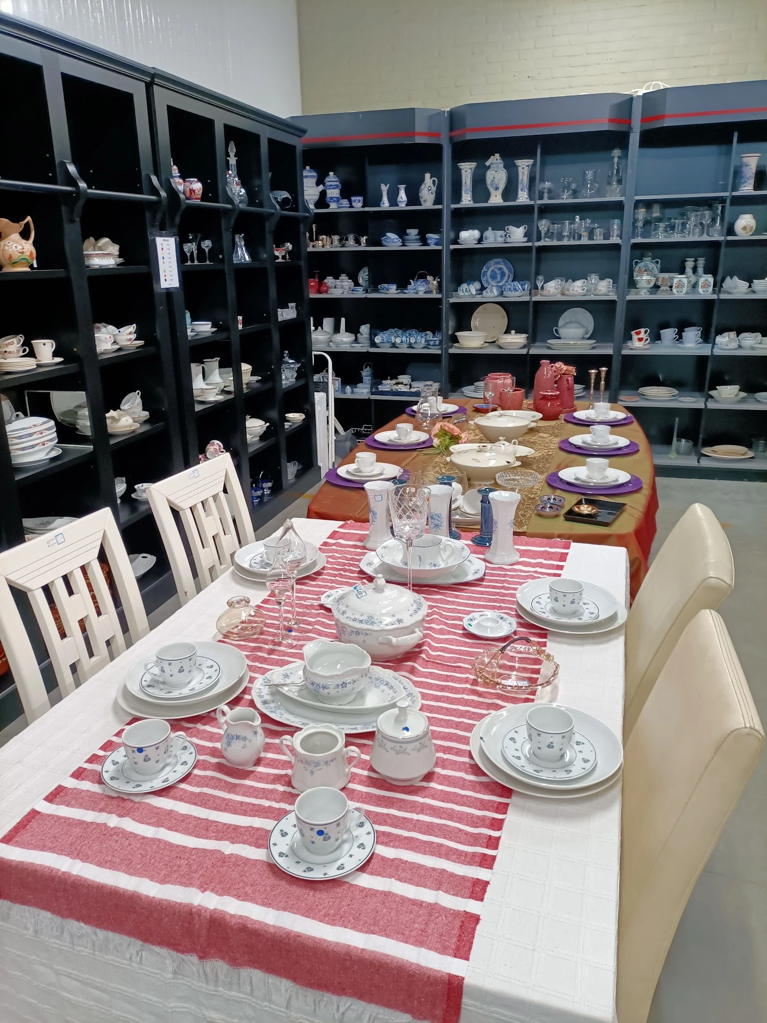 Twee tafels met serviesgoed en tafelschikking; op de achtergrond volle stellingen met aardewerk en servies