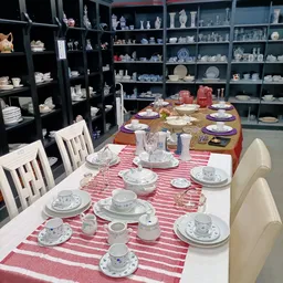Twee tafels met serviesgoed en tafelschikking; op de achtergrond volle stellingen met aardewerk en servies