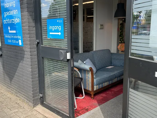 Ingang van kringloopwinkel met zithoek; grijze bank op rood tapijt in entreegebied