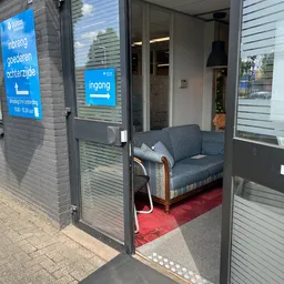 Ingangsruimte van kringloopwinkel met blauwe deur, grijze bank op rode vloerkleed en ingericht interieur