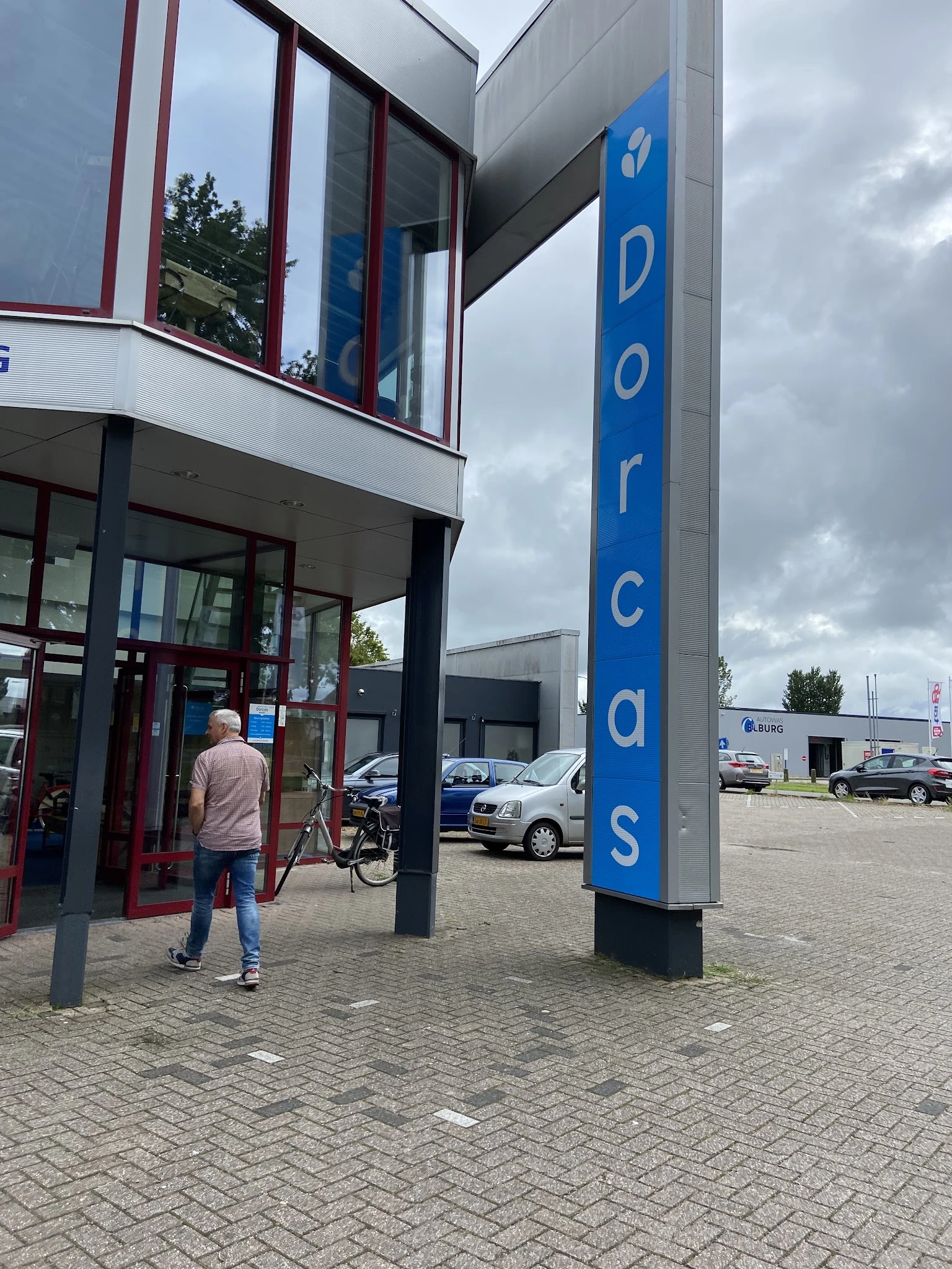 Voorkant van kringloopwinkel met blauwe pilaar met 'Dorcas' tekst en ingang met glazen deuren, persoon met fiets zichtbaar