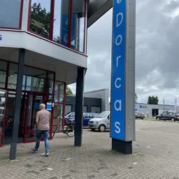 Voorkant van kringloopwinkel met blauwe pilaar met 'Dorcas' tekst en ingang met glazen deuren, persoon met fiets zichtbaar