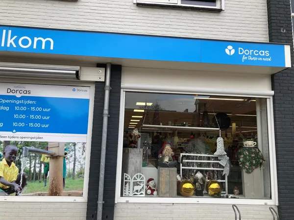 Voorkant van kringloopwinkel Dorcas met blauwe luifel, winkelruit met huisraad en decorstukken, openingstijden zichtbaar