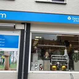 Voorkant van kringloopwinkel Dorcas met blauwe luifel, winkelruit met huisraad en decorstukken, openingstijden zichtbaar