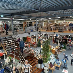 Overzicht van kringloopwinkel met meerdere etages, stellingen met kleding en huishoudartikelen, bezoekers aan het winkelen