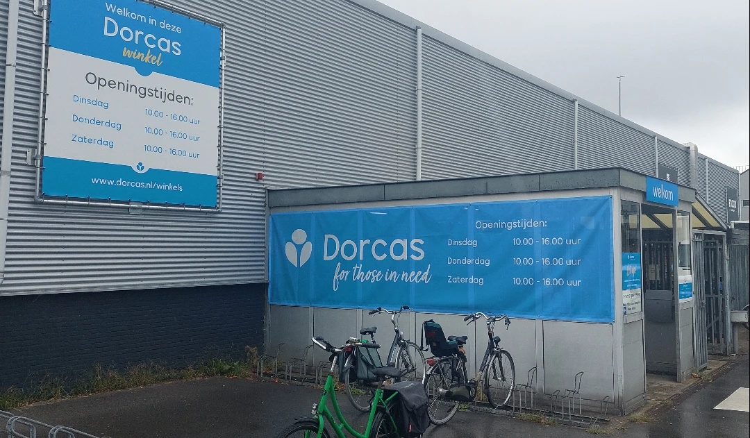Gevel van kringloopwinkel Dorcas met blauwe belettering, openingstijden en logo, fietsenstalling ervoor