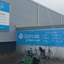 Gevel van kringloopwinkel Dorcas met blauwe belettering, openingstijden en logo, fietsenstalling ervoor