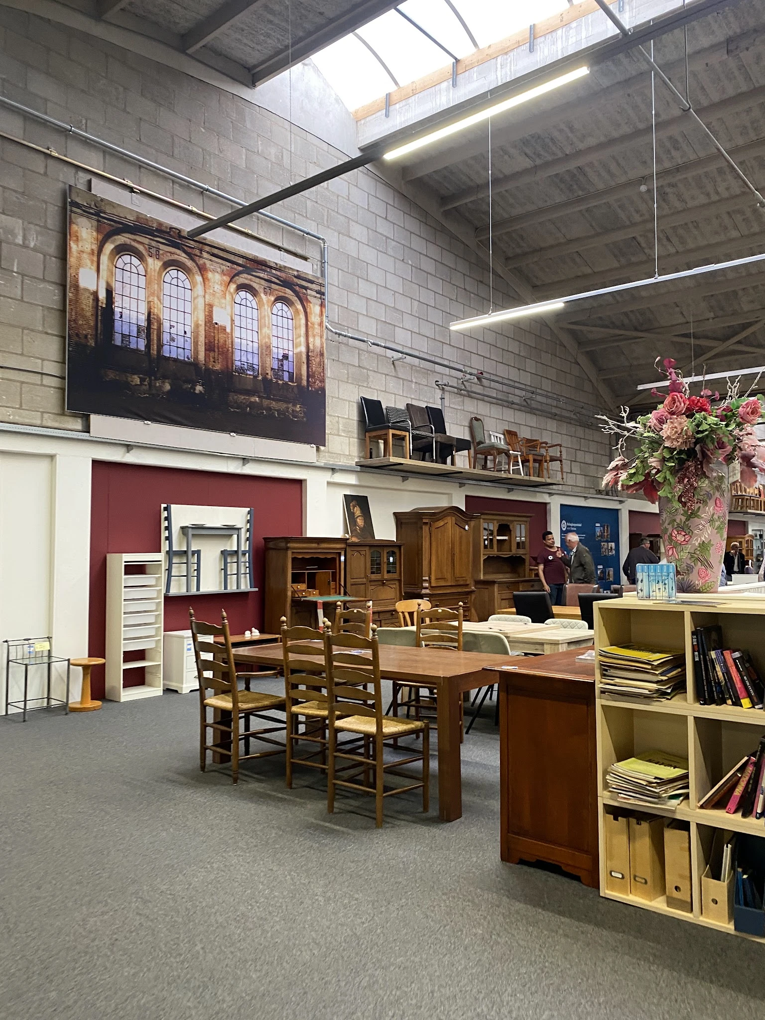 Winkelvloer met houten meubels, boeken, stoelen en diverse spullen uitgestald, rode muur op achtergrond