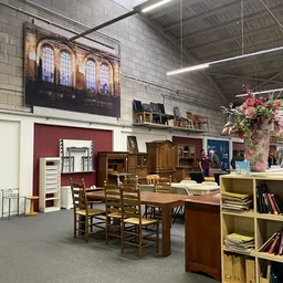 Winkelvloer met houten meubels, boeken, stoelen en diverse spullen uitgestald, rode muur op achtergrond