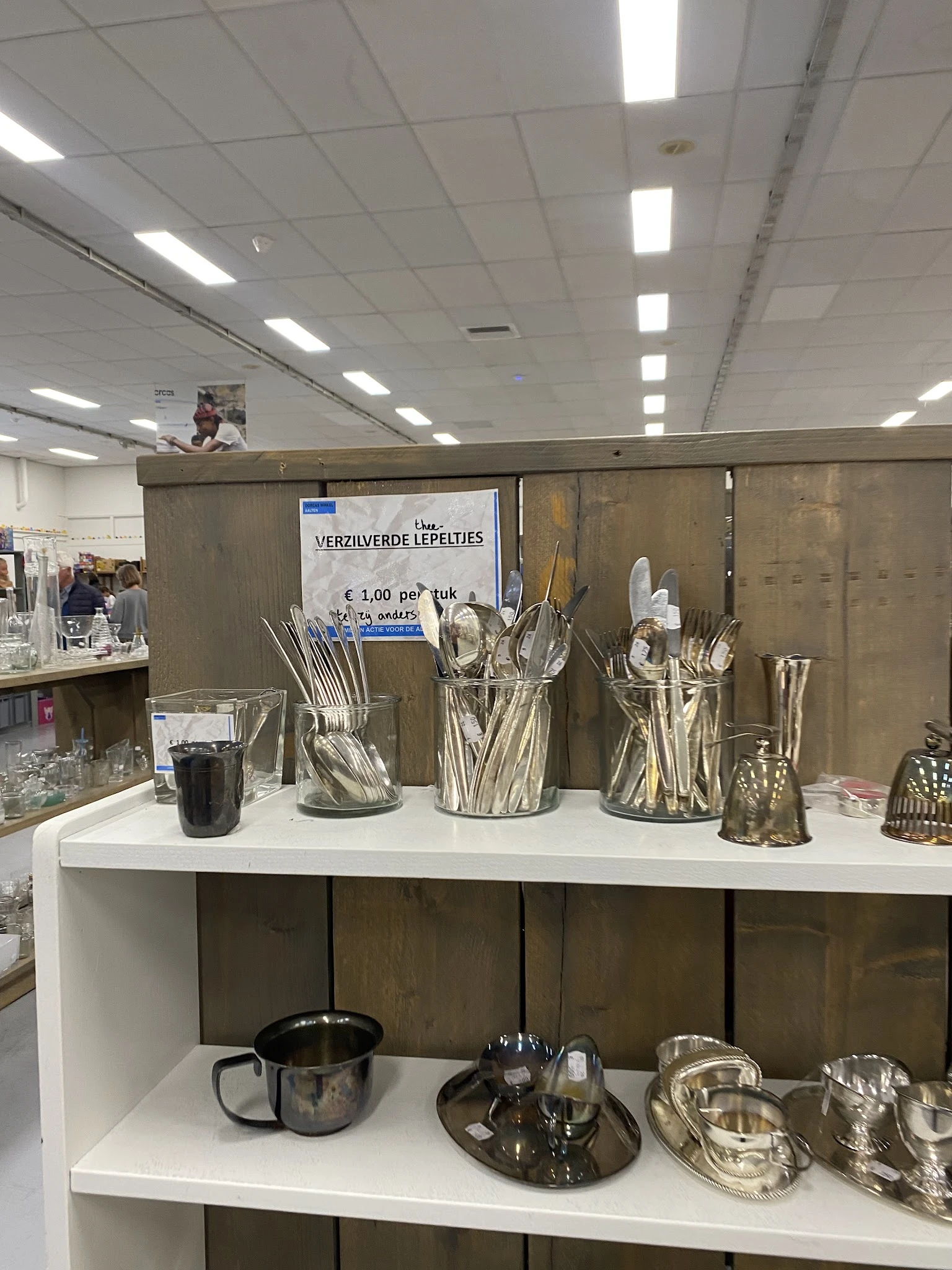 Uitstallingsrek met zilveren bestek in glazen bekers, op witte tafel in kringloopwinkel