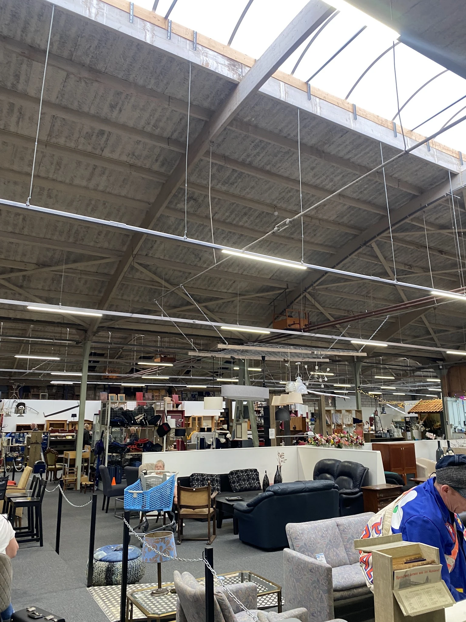 Overzicht van kringloopwinkel met stellingen, meubels en spullen onder industrieel plafond met tl-verlichting