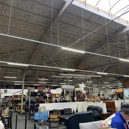 Overzicht van kringloopwinkel met stellingen, meubels en spullen onder industrieel plafond met tl-verlichting