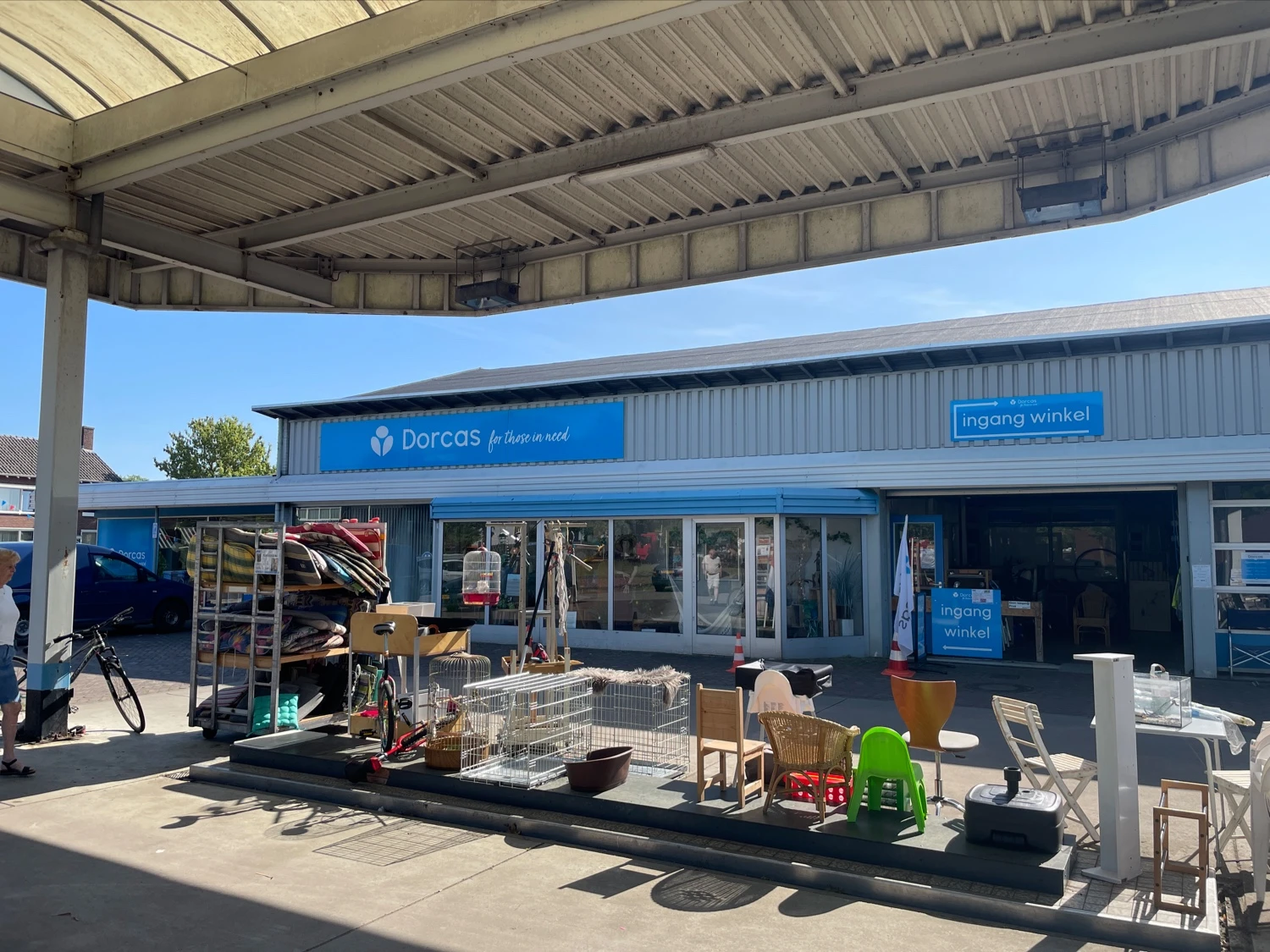 Blauwe gevel van kringloopwinkel met naam Dorcas zichtbaar, onder afdak met spullen uitgestald op het terras