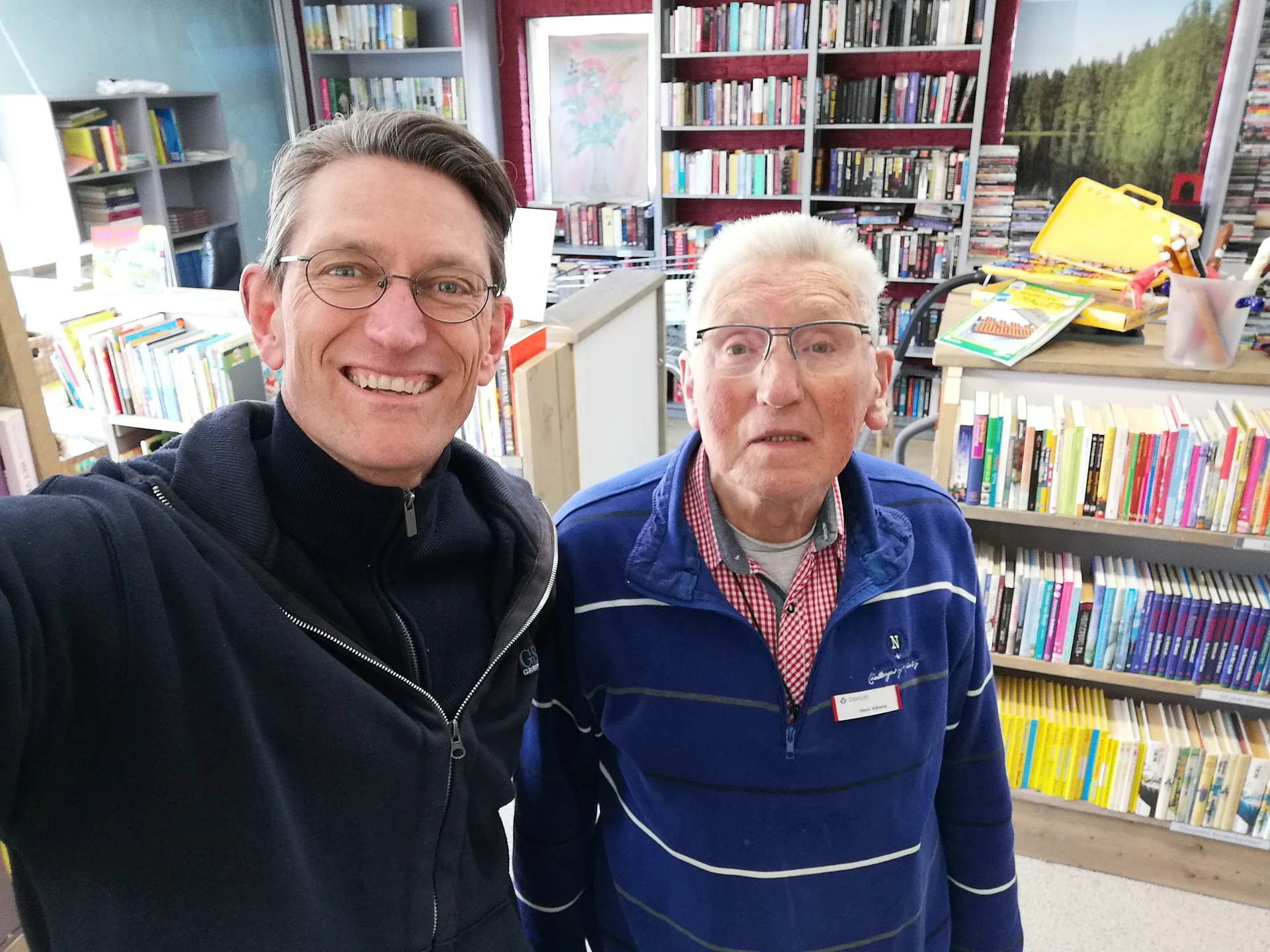 Twee medewerkers poseren voor de camera in een kringloopwinkel met boekenkasten op de achtergrond