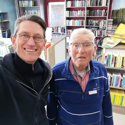 Twee medewerkers poseren voor de camera in een kringloopwinkel met boekenkasten op de achtergrond