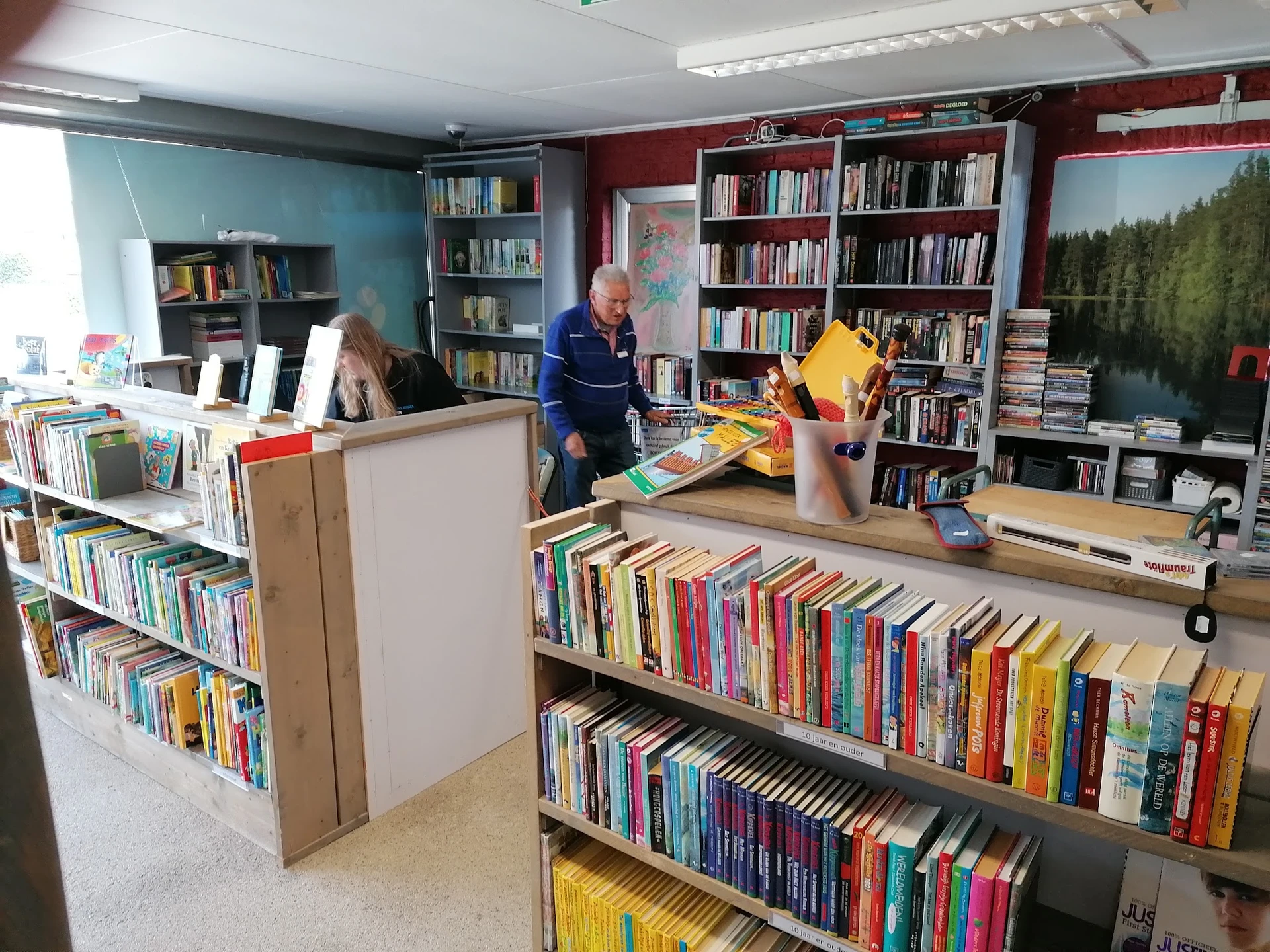 Boekenrekken en stellingen vol met tweedehands boeken in verschillende kleuren, met een baliebediende en klanten in de winkel