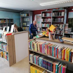 Boekenrekken en stellingen vol met tweedehands boeken in verschillende kleuren, met een baliebediende en klanten in de winkel