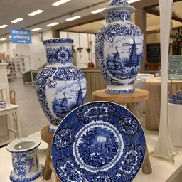 Uitgestalde blauwe en witte Delfts aardewerk: twee vazen en een decoratief bord op houten voetstukken in de winkel