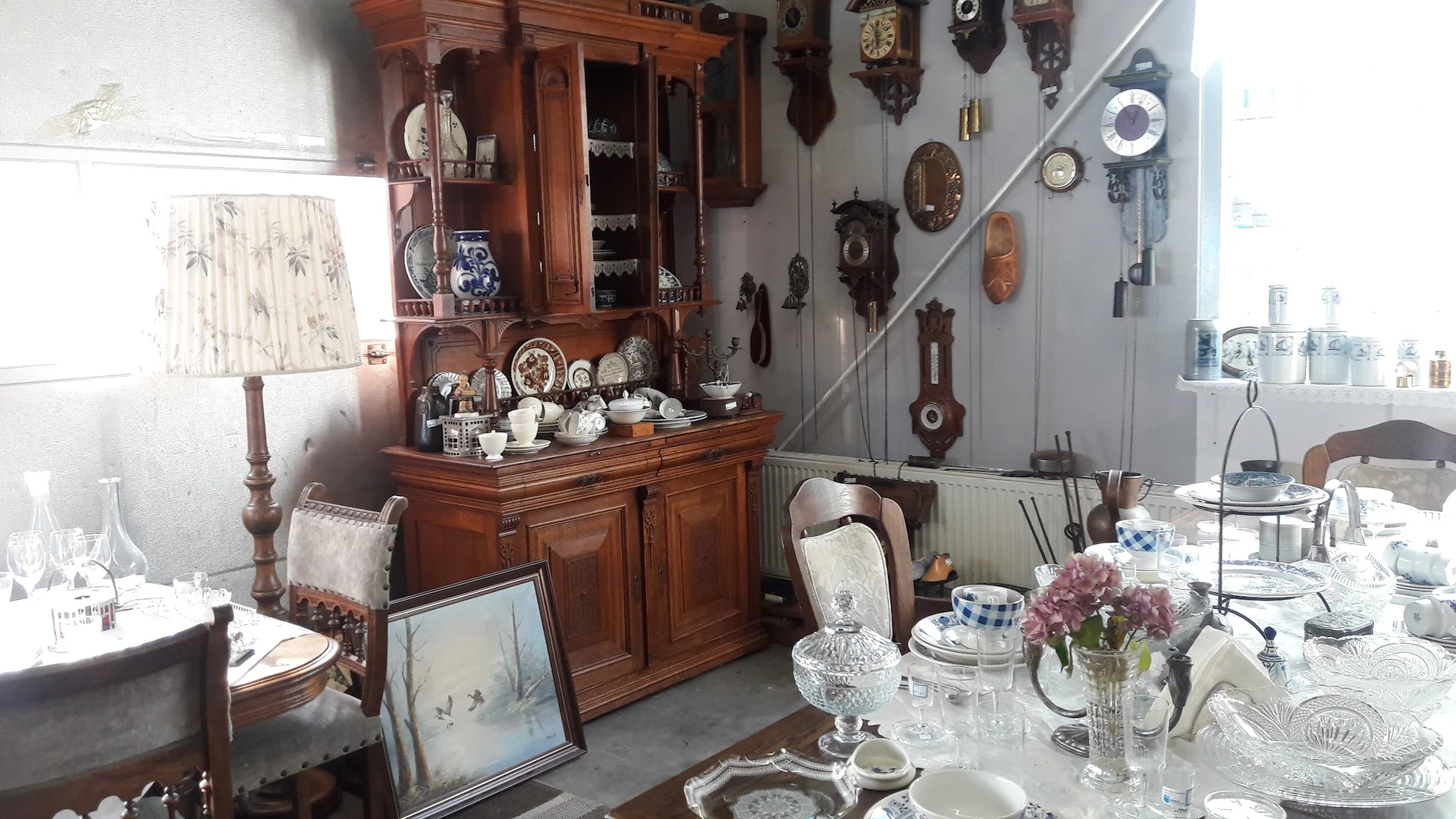 Houten buffetkast met porselein, servies en decoratieve items; aan de muur horloges en diverse antieke spullen