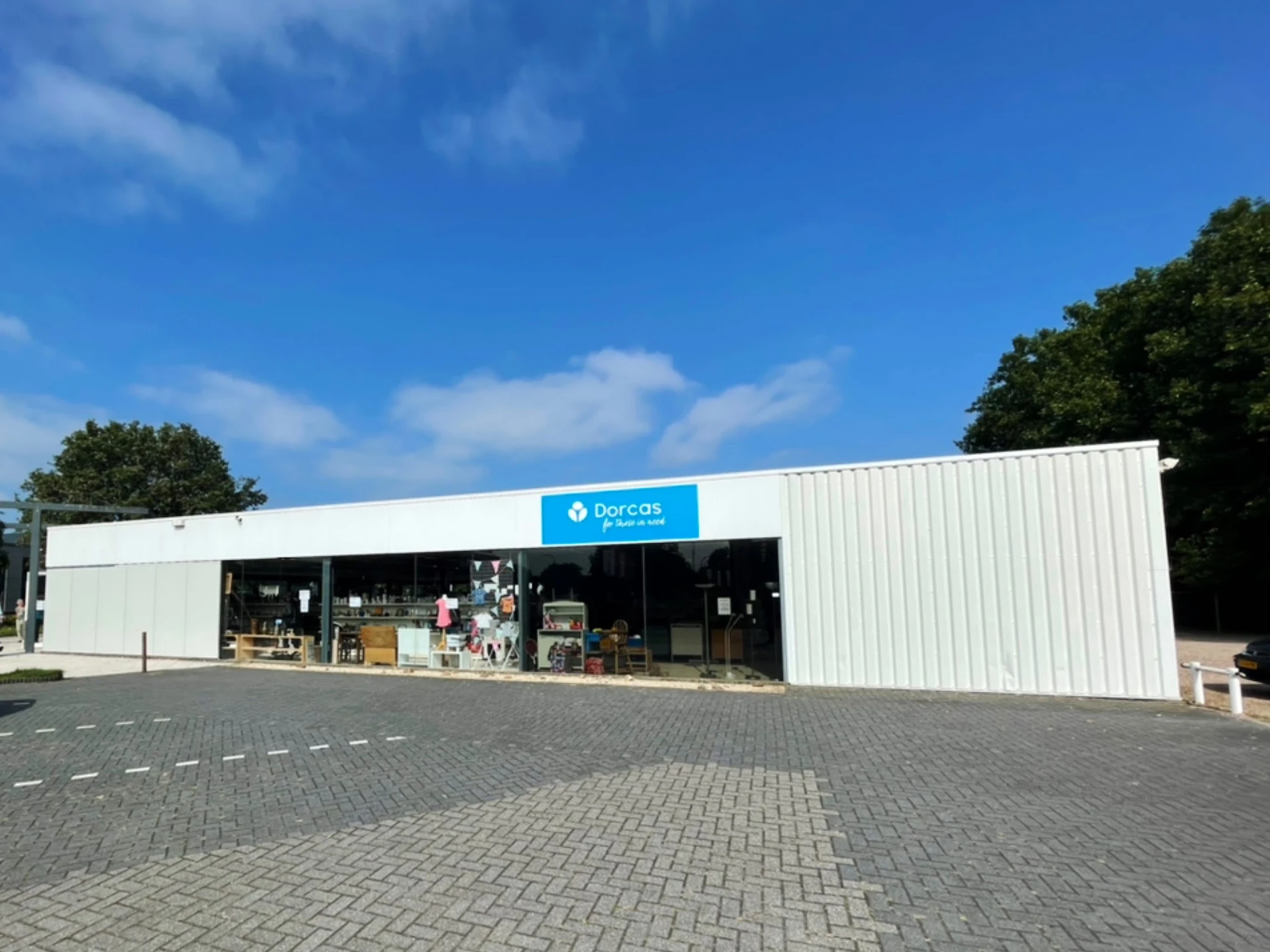 Witte moderne gevel van kringloopwinkel met blauwe Dorcas-logo, parkeerplaats met klinkerwerk, bomen op achtergrond