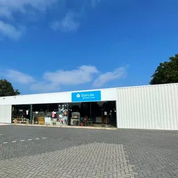 Witte moderne gevel van kringloopwinkel met blauwe Dorcas-logo, parkeerplaats met klinkerwerk, bomen op achtergrond