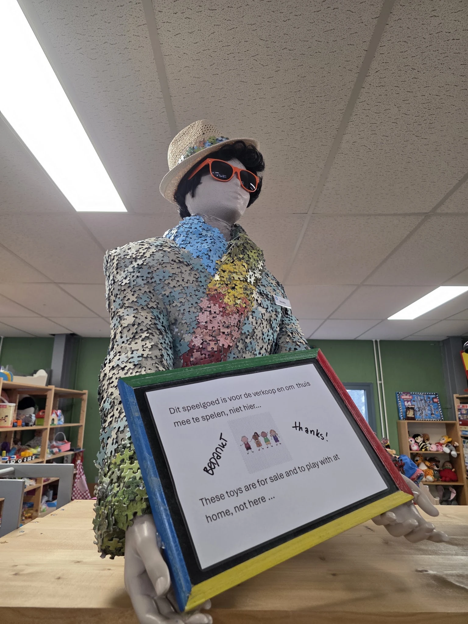 Mannequin gekleed in een zilveren glitterjas met gekleurde vlakken, met hoed en zonnebril, in een winkelinterieur.
