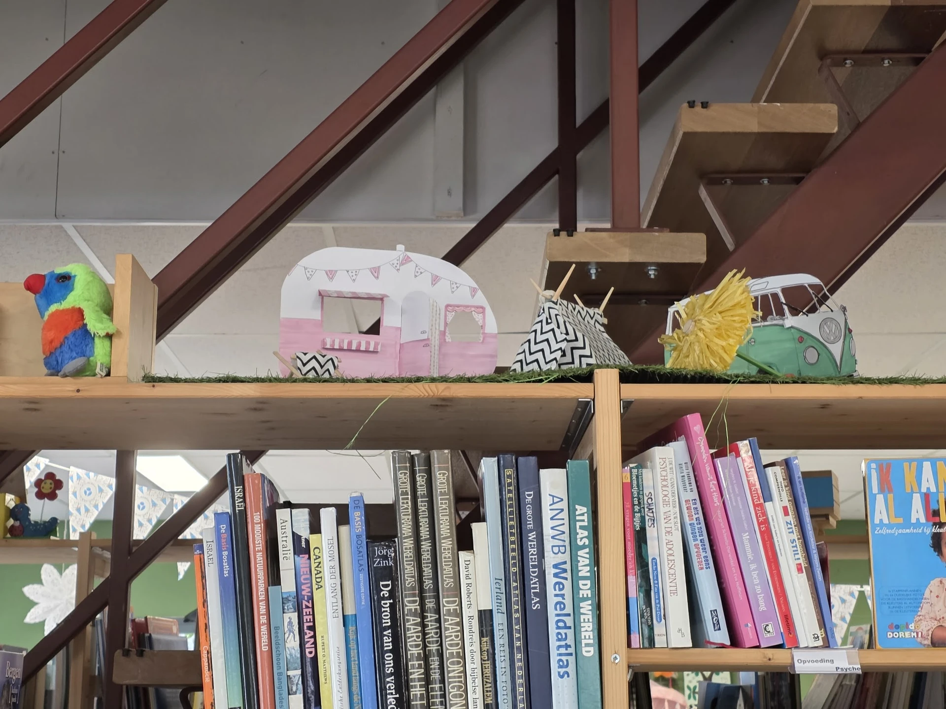 Boeken op een houten plank met daarop decoratieve artikelen: een kleurrijke vogel, roze caravan en groene camper.