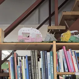 Boeken op een houten plank met daarop decoratieve artikelen: een kleurrijke vogel, roze caravan en groene camper.