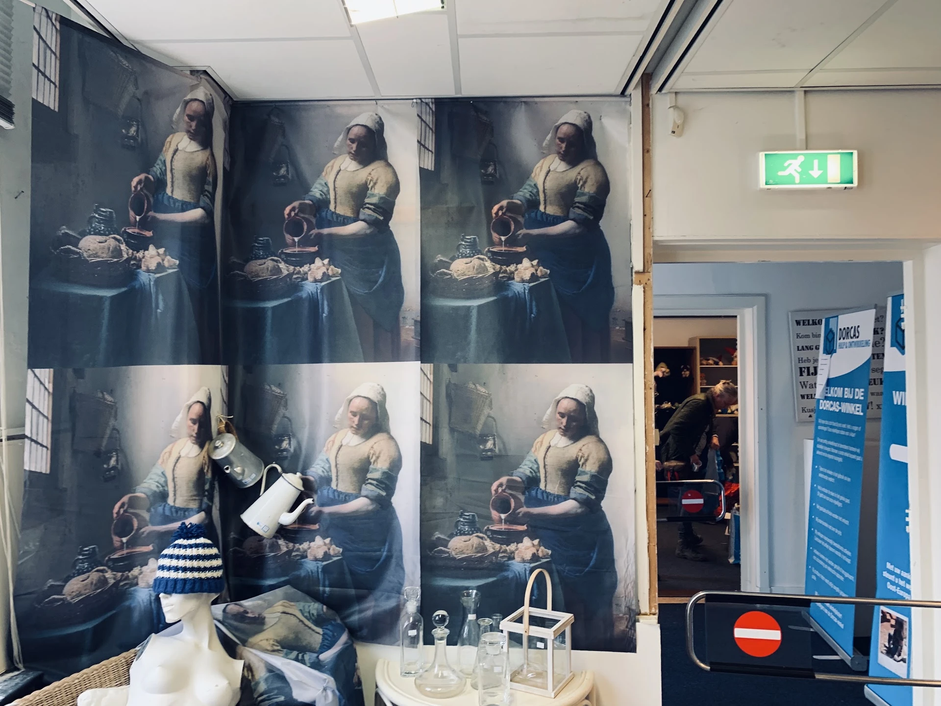 Winkelinterieur met grote kunstdrukken aan de muur en spullen op tafels, groene nooduitgang zichtbaar