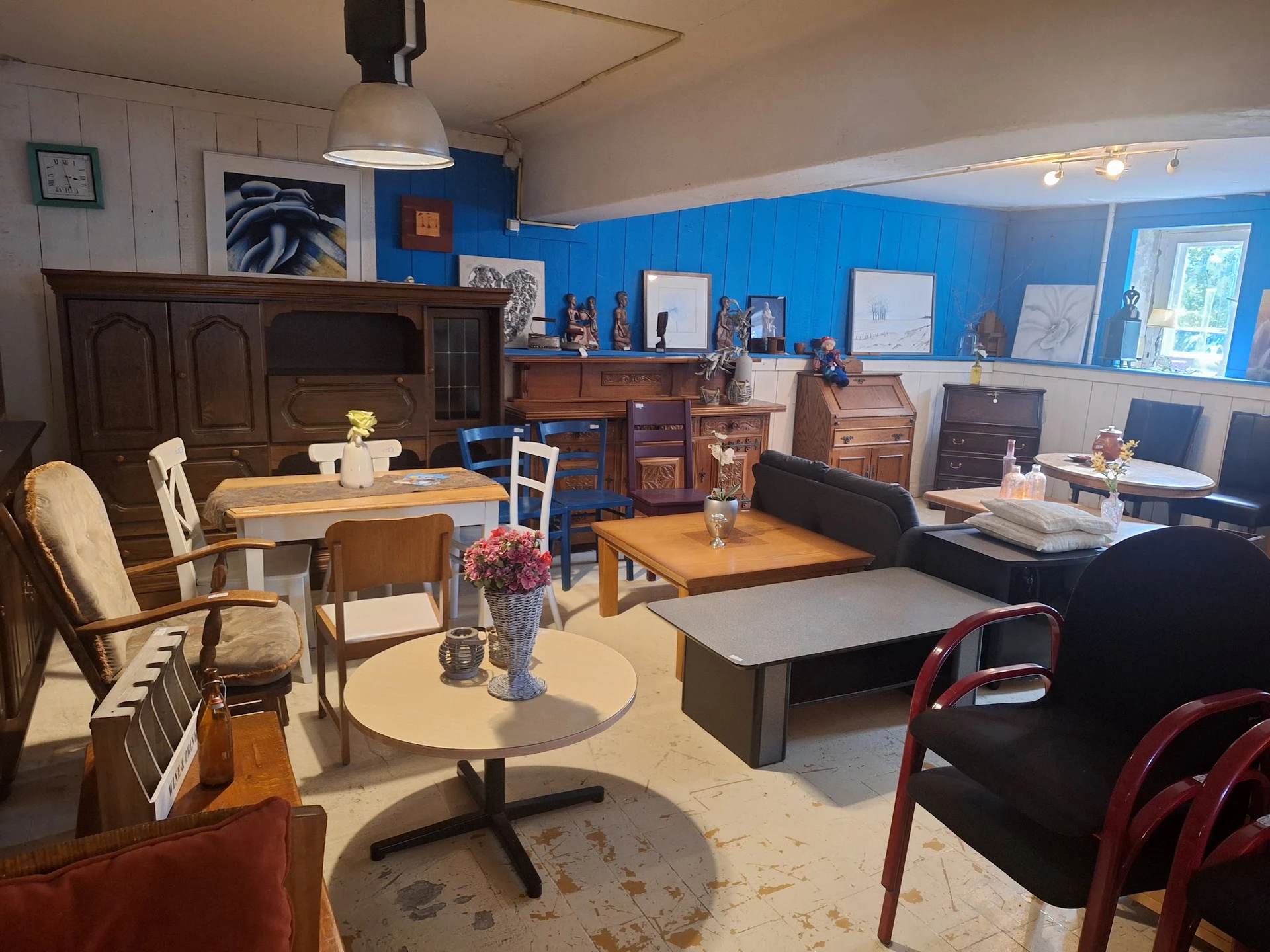 Winkelvloer met diverse meubels, een ronde tafel met bloemen, houten tafels, stoelen en kasten tegen blauwe muur