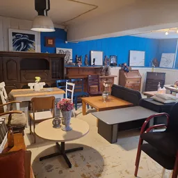 Winkelvloer met diverse meubels, een ronde tafel met bloemen, houten tafels, stoelen en kasten tegen blauwe muur