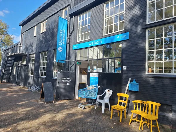 Gevel van kringloopwinkel Dorcas met blauwe luifel, grote ramen en gele stoelen op het terras ervoor