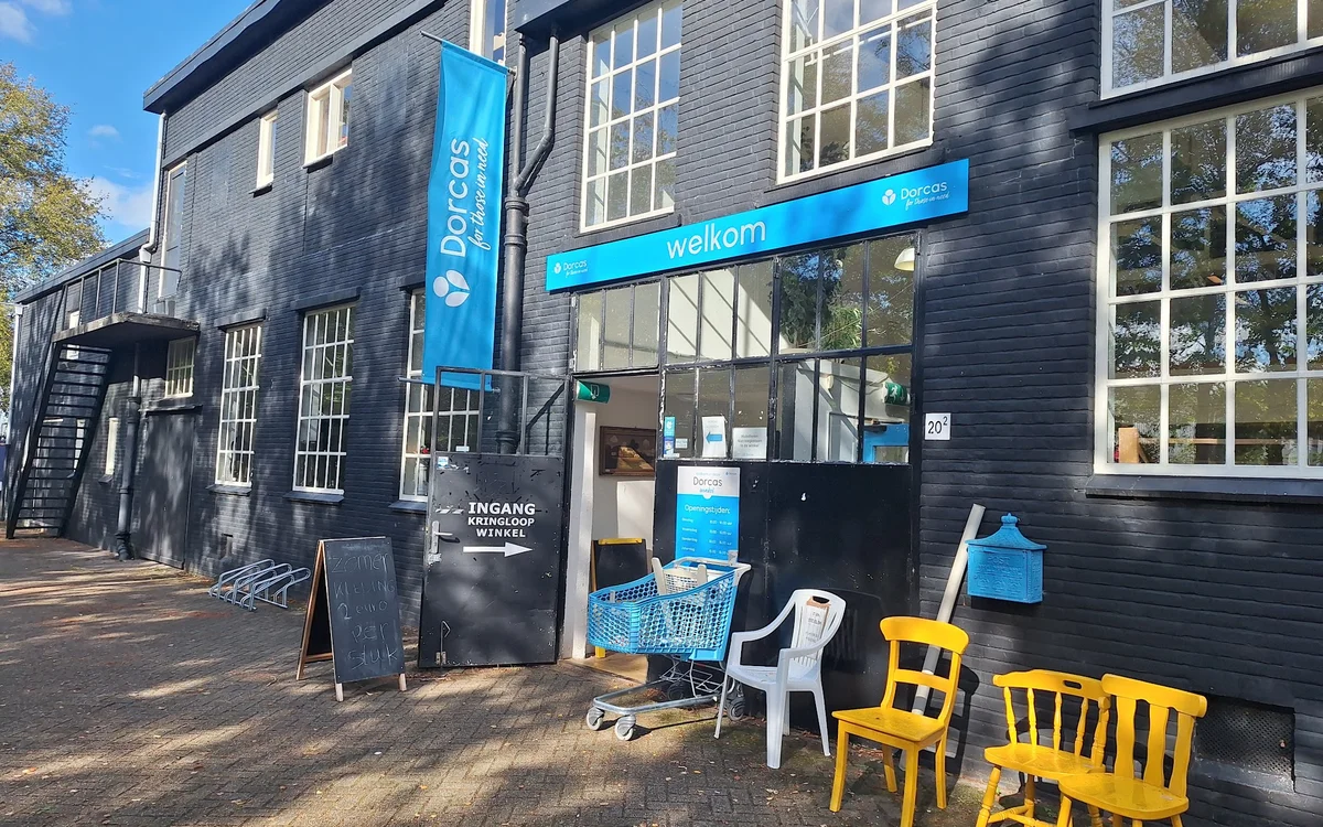 Gevel van kringloopwinkel Dorcas met blauwe luifel, grote ramen en gele stoelen op het terras ervoor