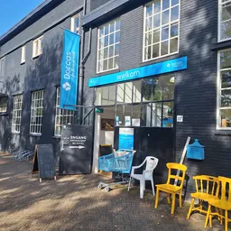 Gevel van kringloopwinkel Dorcas met blauwe luifel, grote ramen en gele stoelen op het terras ervoor