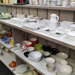 Stellingen vol servies en tafelgerei: witte en gekleurde borden, kommen, bekers en kannen
