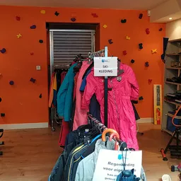 Rekken met tweedehands kleding, jassen en winterjassen in felle kleuren op een oranje muur met klimwandroutes