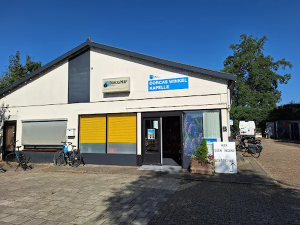 Witte gevel van kringloopwinkel Dorcas Winkel Kapelle met gele deuren, duidelijk zichtbare winkelnaam en logo, fietsen ervoor
