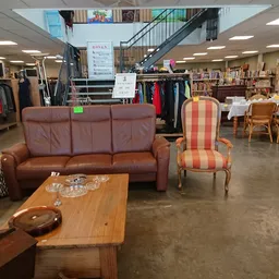 Winkelvloer met bruine leren bank, gestreepte fauteuil en houten tafel; op de achtergrond kledingrekken en boekenkasten