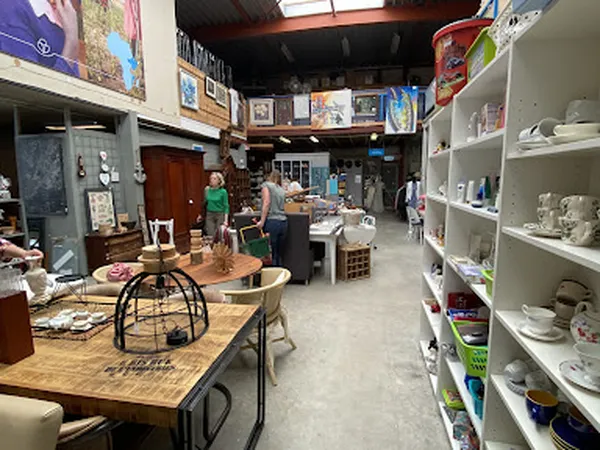 Winkelvloer van kringloopwinkel met houten tafels, witte stellingen met serviesgoed en bezoekers in de winkel